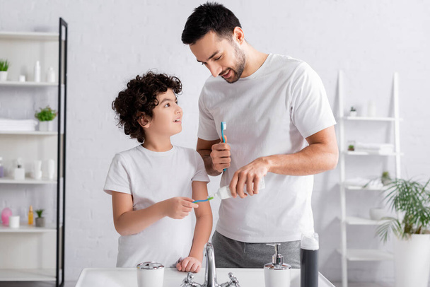 Lächelnder Vater trägt Zahnpasta auf Zahnbürste neben muslimischem Sohn und Waschbecken auf  - Foto, Bild