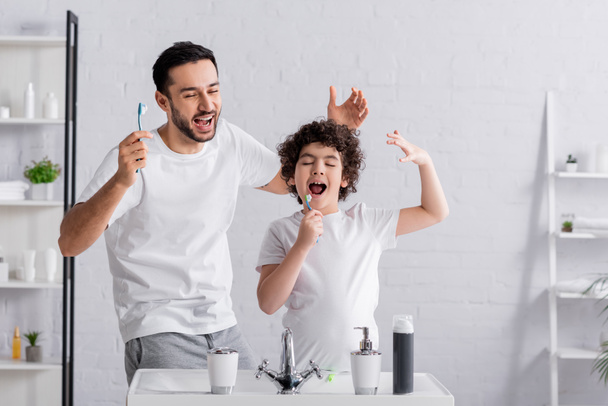Hombre árabe alegre con cepillo de dientes cerca de cantar hijo en el baño  - Foto, Imagen
