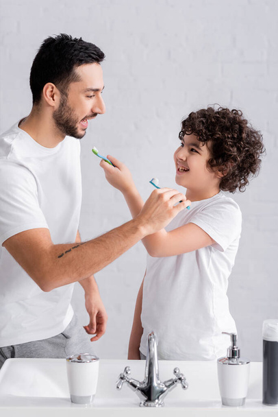 Padre árabe y niño sosteniendo cepillos de dientes cerca del fregadero y el grifo  - Foto, Imagen