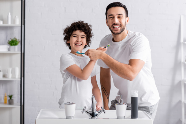 Glücklicher muslimischer Sohn und Vater halten Zahnbürsten in der Hand und lächeln im Badezimmer in die Kamera  - Foto, Bild