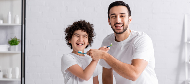Θετικός αραβικός πατέρας και γιος κρατώντας οδοντόβουρτσες και οδοντόκρεμα, πανό  - Φωτογραφία, εικόνα