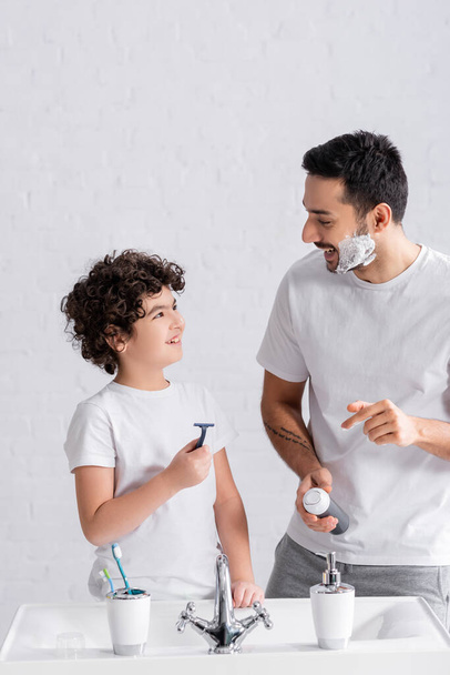 Sonriente hijo musulmán y padre con espuma de afeitar y maquinilla de afeitar en el baño  - Foto, imagen