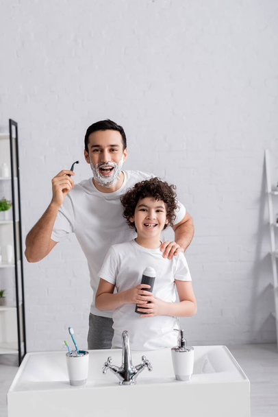 Cheerful arabian man holding razor near son with shaving foam  - Photo, Image