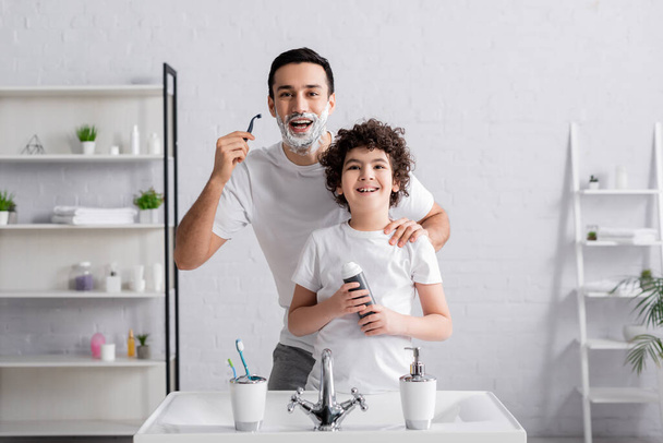 Мусульманин держит бритву рядом с улыбающимся сыном с пеной для бритья в ванной комнате  - Фото, изображение