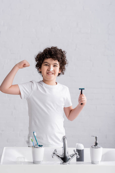 Sonriente chico árabe con espuma de afeitar en la nariz sosteniendo la afeitadora y mostrando músculo en el baño  - Foto, imagen