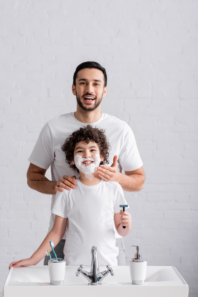 笑顔アラビア人の父適用シェービングフォームの息子の顔に剃刀でバスルーム  - 写真・画像