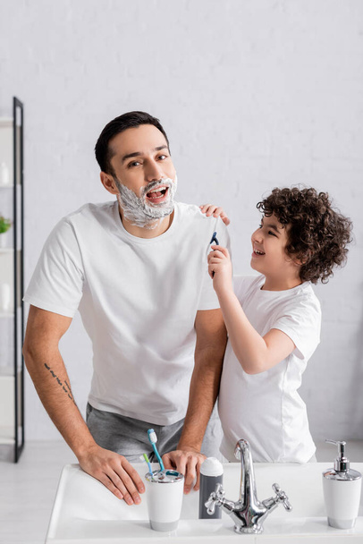 Arabo ragazzo tenendo rasoio vicino sorridente padre in schiuma da barba in bagno  - Foto, immagini