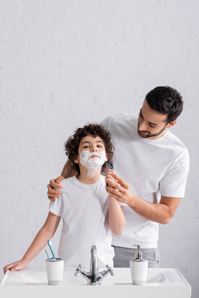 Vater lehrt arabischen Sohn im Rasierschaum mit Rasiermesser im Badezimmer  - Foto, Bild