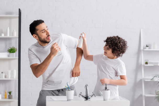 Sonriente hijo árabe sosteniendo desodorante cerca de padre con rociador y fregadero  - Foto, imagen