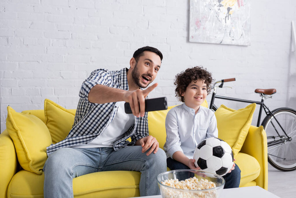 Araber mit Fernbedienung zeigt mit Finger auf Popcorn und Sohn mit Fußball zu Hause  - Foto, Bild