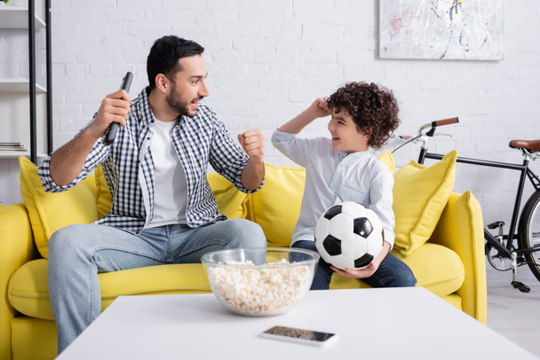 Spannende Arabische man met afstandsbediening op zoek naar zoon met voetbal in de buurt van popcorn en smartphone  - Foto, afbeelding