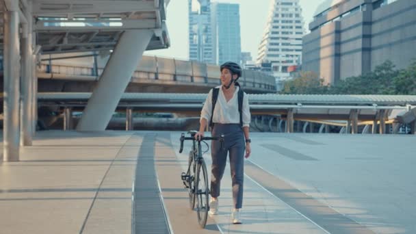 Empresária asiática ir para o trabalho no escritório andar e sorrir usar mochila olhar ao redor tomar bicicleta na rua em torno de construção em uma rua da cidade. Viajar de bicicleta, Viajar de bicicleta, Conceito de comutador de negócios. - Filmagem, Vídeo