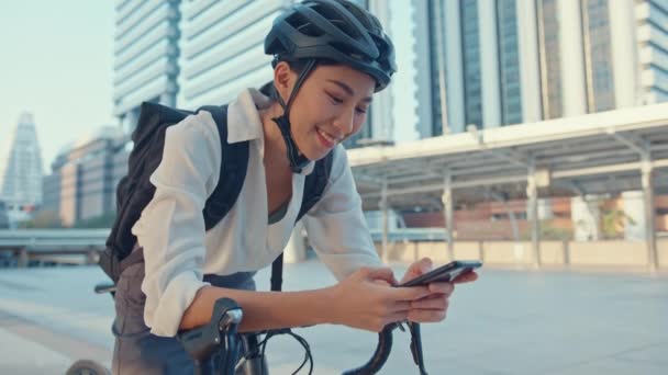 Lächeln asiatische Geschäftsfrau mit Rucksack verwenden Smartphone-Look Kamera in der Stadt stehen auf der Straße mit dem Fahrrad zur Arbeit ins Büro zu gehen. Sport Mädchen verwenden Telefon für die Arbeit. Pendeln zur Arbeit, Berufspendler in der Stadt. - Filmmaterial, Video