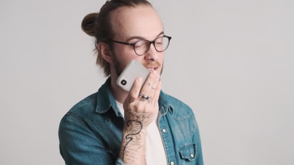Ελκυστικό κομψό γενειοφόρος τύπος σε γυαλιά ηλίου καταγραφή φωνητικό μήνυμα χρησιμοποιώντας smartphone πάνω από λευκό φόντο. Πάντα σε επαφή. - Πλάνα, βίντεο