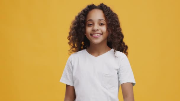 Cute african american curly teen girl gesturing thumbs up and smiling at camera, orange background, slow motion - Footage, Video