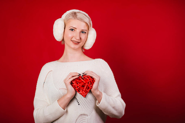 meisje in witte bont koptelefoon in een warme trui op een rode achtergrond. Een blonde vrouw met een hart in haar handen. - Foto, afbeelding