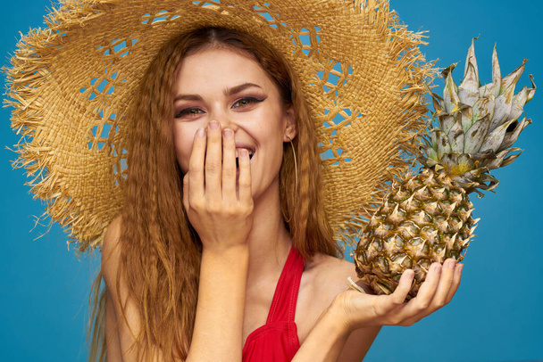 Une femme dans un chapeau de paille avec un ananas dans les mains ou est-ce un fond de fruits exotiques bleu amusant - Photo, image