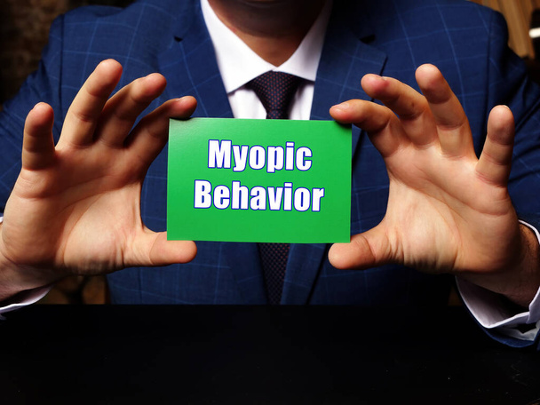 Muž v modrém obleku a ukazující prázdnou zelenou vizitku s psaným textem Myopic Behavior. Rozmazané pozadí. Vodorovné mocku - Fotografie, Obrázek