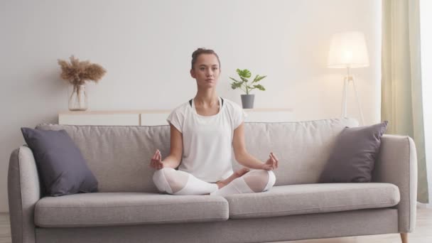 Mladá pozorná žena meditující doma, sedící na gauči v lotosové poloze a dýchající - Záběry, video