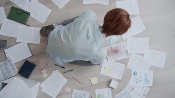 Timelapse plano superior del hombre con el pelo rojo, en camisa de mezclilla y jeans sentados en el suelo, trabajando con documentos, gráficos y diagramas dispuestos a su alrededor, y la escritura en notas adhesivas - Imágenes, Vídeo