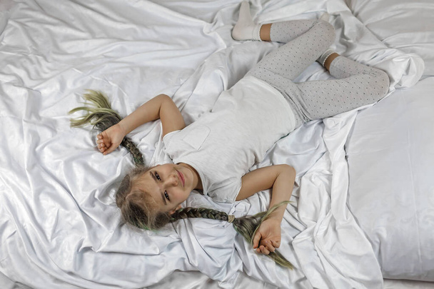 Awaking girl in white clothes with two braids lying on bed in bedroom in the morning, sleep care - Photo, Image