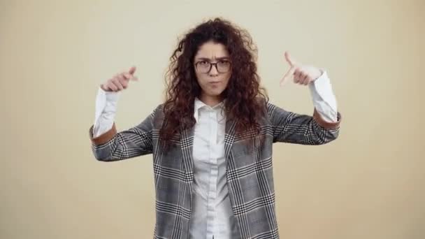 Hey subscribe here, the resentful young woman looks with both fingers down like a gesture. Cretaceous in gray jacket and white shirt, with glasses posing isolated on a beige background in the studio. - Footage, Video