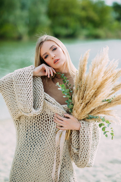 Young blonde woman poses on beach near lake with bouquet of dry flowers in her hands. - Photo, Image