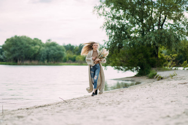 Mladá blondýna žena kráčí podél pobřeží písečné pláže v blízkosti jezera s kyticí suchých květin v jejích rukou. - Fotografie, Obrázek