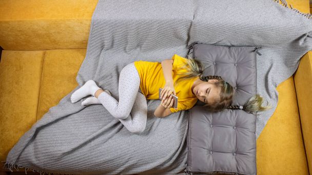 Παιδί σε κίτρινο καναπέ με γκρι κουβέρτα και τη χρήση smartphone για online chat και μακρινή μάθηση - Φωτογραφία, εικόνα