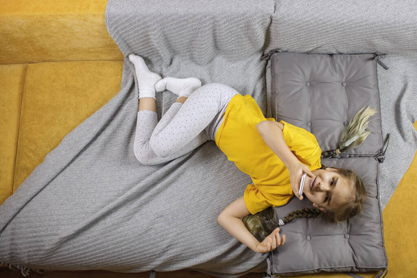 Ребенок на желтом диване с серым одеялом и с помощью смартфона для онлайн чата и дистанционного обучения - Фото, изображение