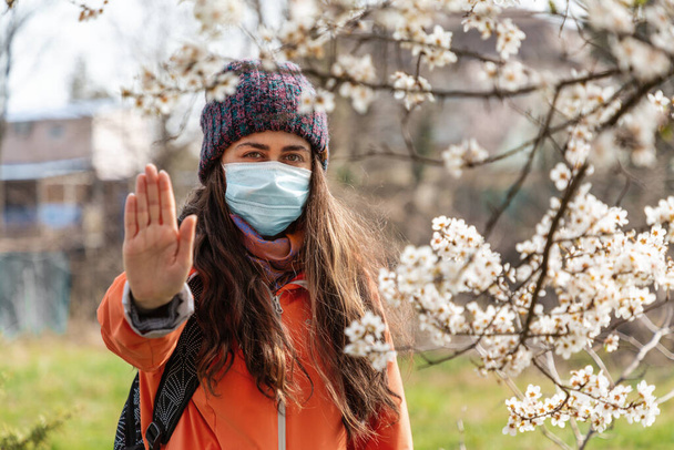 医者のマスクの女性が開花木の近くの通りに立っており、彼女の手で停止記号を示しています。季節性花粉症とウイルス感染症の概念. - 写真・画像