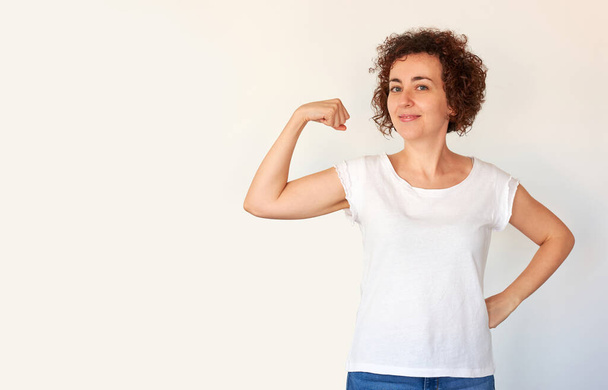 Een gekrulde blanke jonge vrouw pronkt met haar biceps spieren als een teken van succes en gezondheid. Witte achtergrond. Geïsoleerde persoon. - Foto, afbeelding