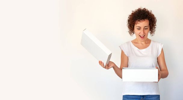 Eine lockige junge Frau öffnet einen weißen Schuhkarton, den sie in einem Online-Shop bestellt hat. Sie liefert Pakete schnell nach Hause. Jubiläumsüberraschung. Weißer Hintergrund. Weißes T-Shirt und Jeans. - Foto, Bild