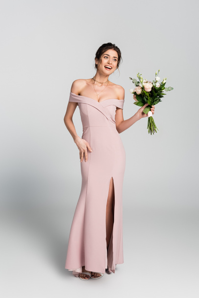 full length view of excited woman in long wedding dress holding bouquet and looking away on grey - Photo, Image
