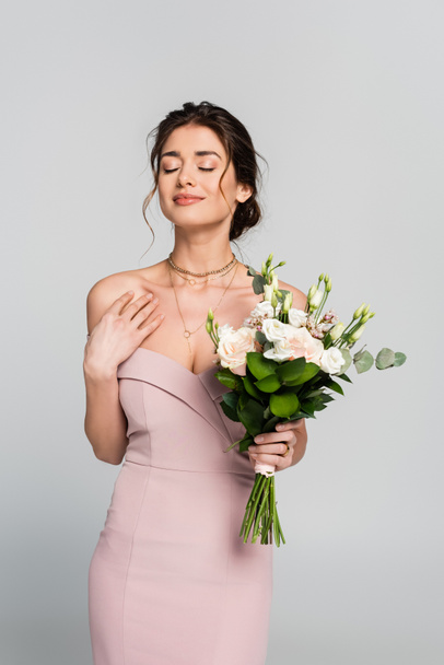 smiling woman holding wedding bouquet while standing with closed eyes isolated on grey - Photo, Image