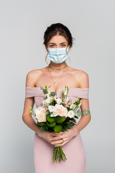 junge Verlobte in medizinischer Maske hält Brautstrauß isoliert auf grau - Foto, Bild