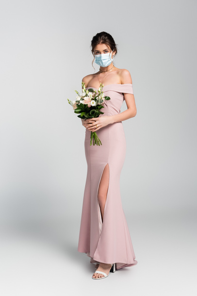 vista completa della sposa in maschera medica in posa con bouquet da sposa su grigio - Foto, immagini