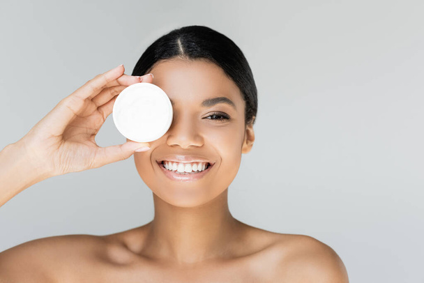 happy african american woman covering eye while holding container with face cream isolated on grey - Photo, Image