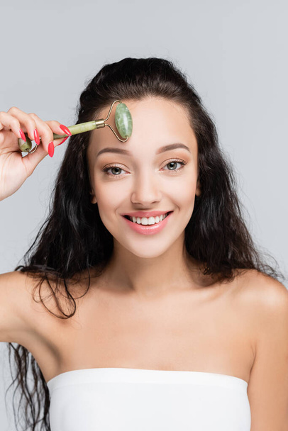 feliz jovem mulher usando jade rolo enquanto massagear rosto isolado em cinza - Foto, Imagem