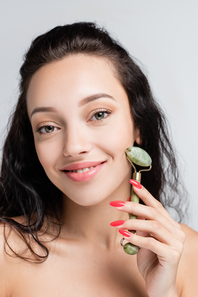 smiling young woman using jade roller while massaging face isolated on grey - Photo, Image
