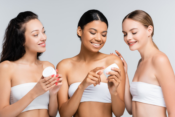vidám, többnemzetiségű, csupasz vállú nők, akik szürke alapon izolált kozmetikai krémmel tartják a konténereket - Fotó, kép