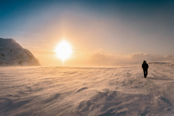 Scenic beeld van grote zonsondergang halo in de winter bergen, sterke sneeuw drift wind. Een persoon in warme zwarte sneeuwscooter algemeen en bril loopt tegen sterke wind. Lapland, Zweden - Foto, afbeelding