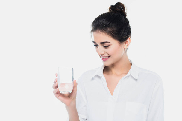 Νεαρή γυναίκα χαμογελά και κοιτάζει ένα ποτήρι νερό απομονωμένο στο λευκό  - Φωτογραφία, εικόνα