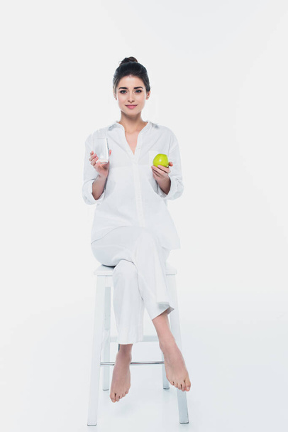 Giovane donna con mela e bicchiere d'acqua sulla sedia su sfondo bianco  - Foto, immagini