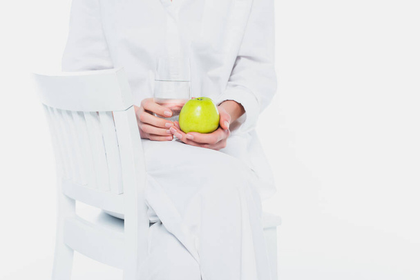 Widok kobiety ubranej w białe ubrania trzymającej jabłko i szklankę wody na krześle odizolowanym na biało  - Zdjęcie, obraz