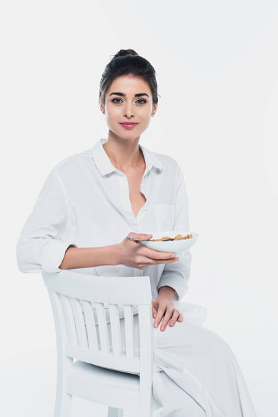 Brunette femme tenant bol de céréales sur chaise isolé sur blanc  - Photo, image