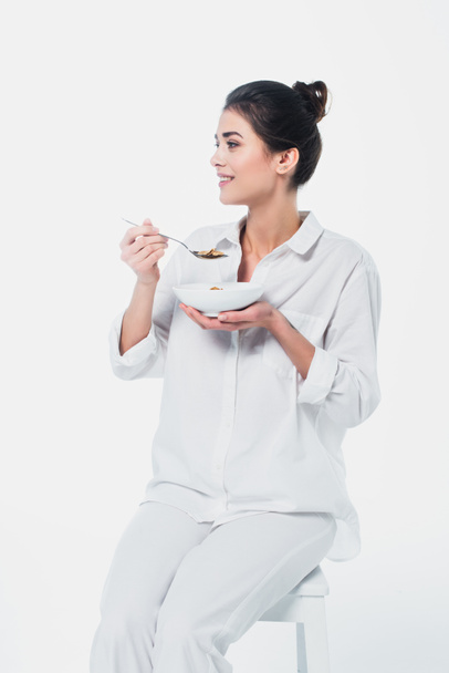 Mulher sorrindo segurando tigela de cereais e colher na cadeira isolada no branco  - Foto, Imagem