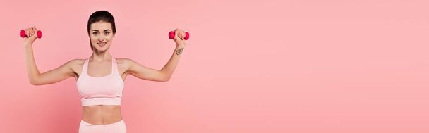 Esmer sporcu kadın, dambıllarla egzersiz yapıyor ve pembe pankartta izole edilmiş kameraya gülümsüyor.  - Fotoğraf, Görsel