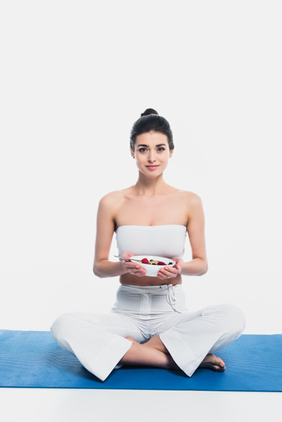Mujer con piernas cruzadas sosteniendo tazón de cereales con bayas en la esterilla de fitness aislado en blanco  - Foto, imagen
