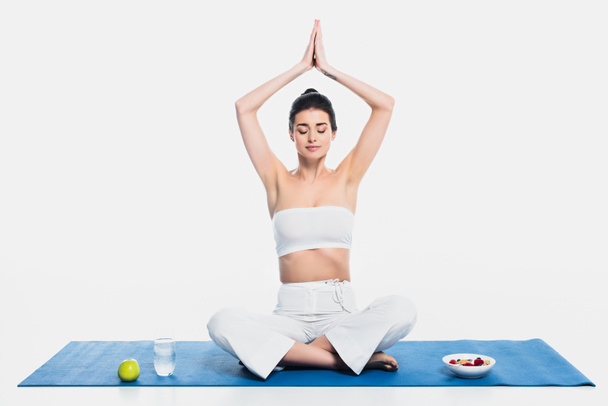 Femme brune méditant près de céréales, pomme et eau sur tapis de fitness sur fond blanc  - Photo, image